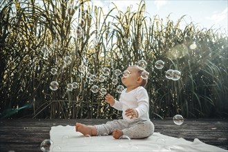 A baby on a blanket surrounded by sunlight, playing with soap bubbles