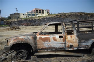 Destroyed lorry in a devastated landscape with burning trees and a building in the background,