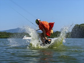 Young casual man with flapping shirt on wakeboard jumping into the lake, water ski and wakepark,