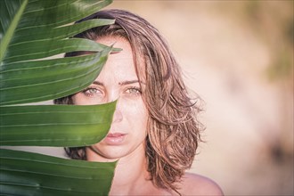 A woman looks out from behind a large green leaf, her face half in the sunlight