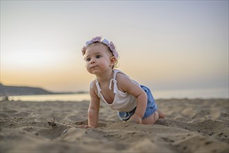 A baby in summer clothes crawls on the beach at sunset