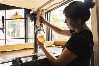 Profile silhouette of a woman serving draft beer in a bar with large windows during the day. The