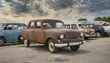 An old, rusty car in a junkyard with other dilapidated vehicles under a cloudy sky, symbol photo,