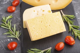 Various types of cheese with rosemary and tomatoes on black slate board on a black concrete