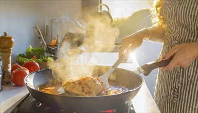 A person is cooking chicken in a pan on a stove. The pan is filled with a red sauce and the chicken