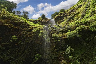 High waterfall falling from green cliffs with lush vegetation and blue sky, Cascata do Grená