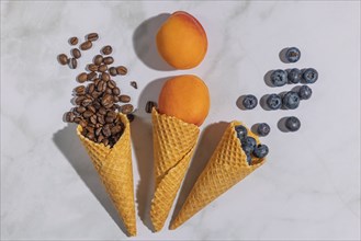 Ice cream cones filled with coffee beans, apricots and blueberries on a marble background