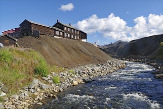 A rushing river next to old buildings on a hill under a sky with white clouds, old mine,