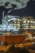 Night scene of an illuminated factory with steaming chimneys and parked vehicles, Mannheim,