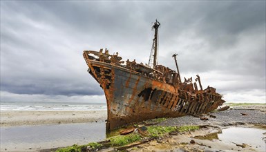 A dilapidated, rusty shipwreck stands against a dark, threatening sky on the coast, symbol photo,