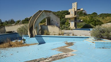 Abandoned swimming pool with concrete ruins and graffiti, surrounded by trees on a sunny day, Lost