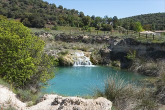 A small waterfall plunges into a turquoise-coloured lake, surrounded by dense meadows and hilly