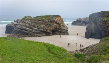 People walking on the beach between rocky cliffs and green grass under a cloudy sky, Praia das