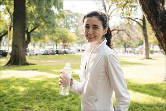 Middle aged woman smiles and looks at camera with water bottle in hand, after workout in park