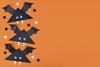 Cute paper vampire bats with funny googly eyes on side of orange Halloween background with empty