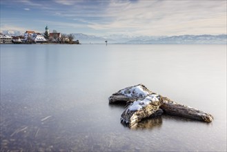 Wooden trunk and picturesque church of St George on the lakeshore in front of snow-covered Pfänder