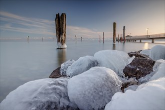 Frozen stones on the shore of a calm lake with posts in the background and soft evening light,