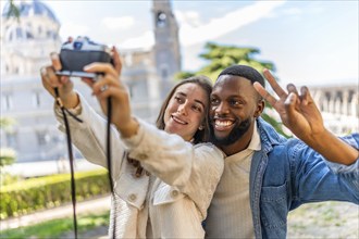 Multi-ethnic couple gesturing success while taking selfie with digital mirrorless camera in a park