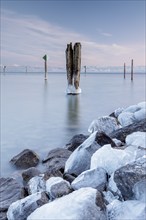 A wooden pile and ice-covered stones on a calm body of water under a blue sky in winter, harbour,