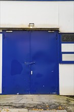 Blue industrial swing gate on a white building with security lighting, with signs of age and