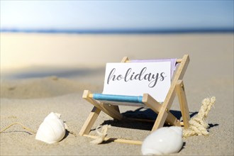 HOLIDAYS text on paper greeting card on background of beach chair lounge summer vacation decor.