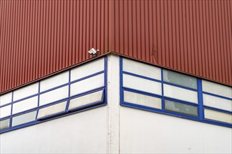 A single surveillance camera is mounted on the corner of a red industrial building with blue-framed