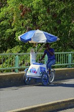 A street vendor selling ice cream rides his bicycle under a blue umbrella in a tropical