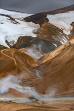 Bridge over steaming stream between colourful rhyolite mountains with snowfields, Hveradalir