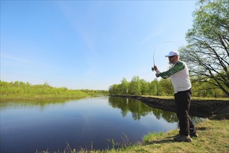 Fisherman with a spinning rod catching fish on a river at sunny summer day with green trees at