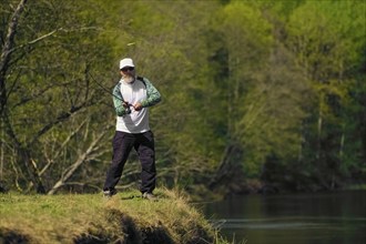 Fisherman throwing the bait into river standing on river bank in sunny day with green forest in