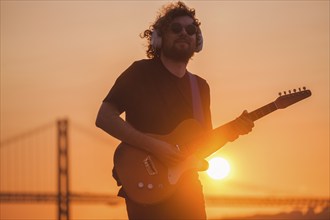 Hipster street musician in black playing electric guitar in street outdoors on sunset