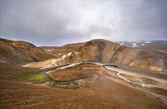 Bridge over steaming stream between colourful rhyolite mountains with snowfields, Hveradalir