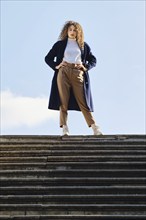 Serious woman in blue wool trench coat stands on stairs in sunny autumn day