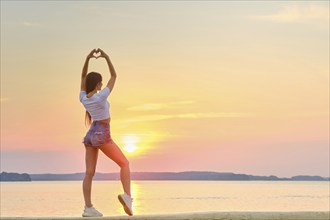 Woman raised her hands up and folded her palms in shape of heart against backdrop of sunset