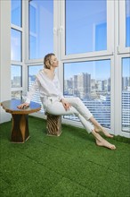 Barefoot young woman sits on the balcony of her new apartment and admires the view of the city from