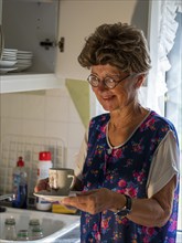 Granny with old smock apron, glasses and wig at the kitchen window in the kitchen, Germany, Europe