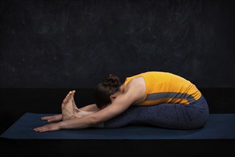 Woman doing Ashtanga Vinyasa Yoga back bending asana Paschimottanasana, seated forward bend on dark