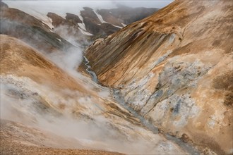 Steaming stream between colourful rhyolite mountains and snowfields, Hveradalir geothermal area,