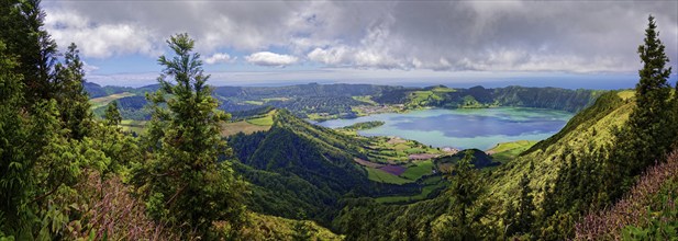 Wide panorama of the crater lake Lagoa Azul surrounded by lush green hills and clouds in the sky,