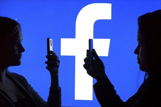 Silhouettes of two woman with smartphones in front of Facebook logo, 12.01.2016