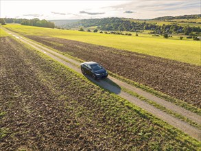 A black car drives along a country lane, surrounded by vast fields under a cloudy sky, Carsharing,