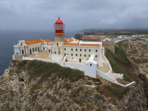 A red lighthouse on a rocky cliff, surrounded by white buildings, with a dramatic view of the