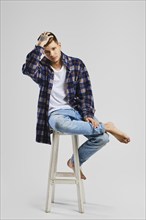 Full length studio portrait of young cocky man in shirt and jeans sitting on tall wooden chair