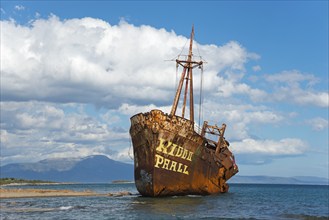 A rusty shipwreck protrudes from the water against a picturesque coastal backdrop, Dimitrios