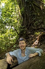 Young man with headlamp, tourist in tropical rainforest, climbing out of the root of a hollow tree,