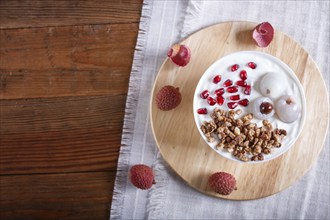 Greek yogurt with lychee, pomegranate seeds and granola in a white plate on a brown wooden