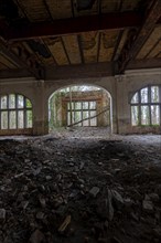 Interior view of a dilapidated building with arched windows and debris on the floor,