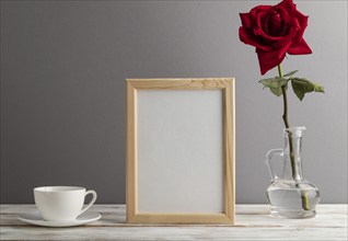 Beige wooden frame mockup with red rose in glass and white coffee cup on gray paper background.