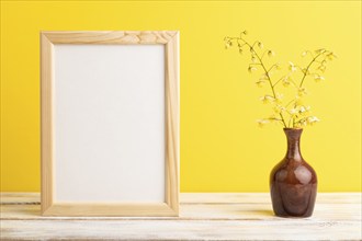 Wooden frame with yellow barrenwort flowers in ceramic vase on yellow pastel background. side view,