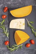 Various types of cheese with rosemary and tomatoes on black slate board on a black concrete